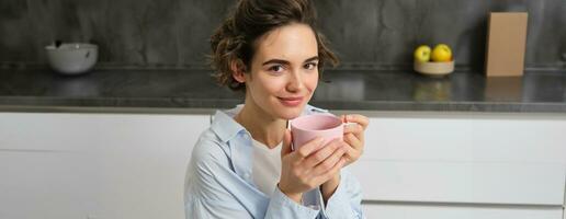 gelukkig ochtenden. portret van gelukkig brunette vrouw, drankjes kop van koffie in haar keuken en lachend, knus en warm begin van de dag met cuppa foto