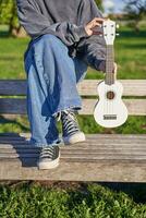 bijgesneden schot van jong meisje in sportschoenen en jeans, handen Holding ukulele musical instrument terwijl ze zit Aan bank in groen zonnig park foto