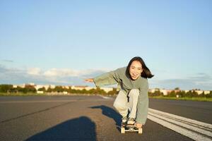 schaatser meisje rijden Aan skateboard, staand Aan haar longboard en lachend, rijden kruiser Aan een leeg straat naar de zon foto