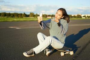 mooi Koreaans meisje duurt selfie Aan smartphone, duurt foto met haar skateboard, geniet zonnig dag buitenshuis