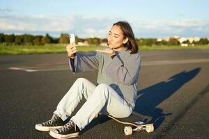 gelukkig Aziatisch meisje zit Aan skateboard, duurt selfie met longboard, maakt schattig gezichten, zonnig dag buitenshuis foto