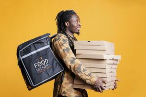 Afrikaanse Amerikaans arbeider draag- stack van pizza, levering snel voedsel bestellingen naar klanten gedurende lunch tijd. restaurant koerier staand in studio met geel achtergrond. voedsel levering en onderhoud foto