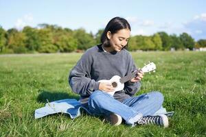 mooi Aziatisch meisje zittend in park, spelen ukulele en zingen, ontspannende buitenshuis Aan zonnig voorjaar dag foto