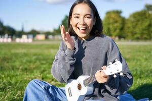 portret van schattig Aziatisch meisje met musical instrument. jong vrouw met verrast gezicht, Holding ukulele en zittend in park Aan deken foto