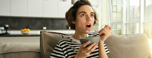 dichtbij omhoog portret van vrouw aan het eten in voorkant van TV, op zoek versteld staan en met interesseren Bij scherm, aan het eten ontbijt, Holding kom van granen en lepel, zittend Aan sofa gefascineerd foto