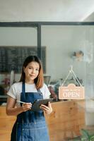 jong vrouw hangende een Welkom teken in voorkant van een koffie winkel. mooi serveerster of gastvrouw Holding een tablet voorbereidingen treffen in een restaurant. foto