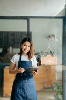 jong vrouw hangende een Welkom teken in voorkant van een koffie winkel. mooi serveerster of gastvrouw Holding een tablet voorbereidingen treffen in een restaurant. foto