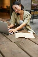 portret van jong vrouw pratend Aan mobiel telefoon terwijl typen, gebruik makend van laptop, werken van een afstand van buiten de deur cafe, aan het doen huiswerk en roeping foto