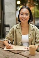 verticaal schot van jong Aziatisch leerling, meisje in bril Holding pen, maken notities, schrijven in notitieboekje en drinken koffie in cafe foto