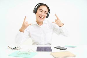 gelukkig en opgewonden Koreaans vrouw in koptelefoon, manager zit Bij bureau in kantoor met digitaal tablet en werk documenten, shows rots n rollen teken, luistert muziek- foto