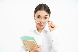 portret van Koreaans vrouw docent, looks onder bril met sceptisch gezicht uitdrukking, Holding notitieboekjes, wit achtergrond foto