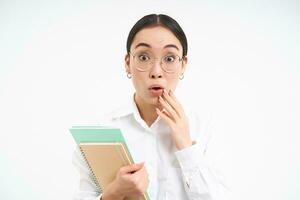 portret van zakelijke vrouw, Aziatisch vrouw in bril, houdt notitieboekje en looks verrast, versteld staan door iets, staat over- wit achtergrond foto