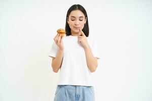 bakkerij en snoepgoed. gelukkig Aziatisch vrouw op zoek Bij smakelijk koekje, aan het eten gebakje, wit achtergrond foto