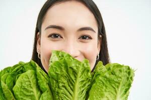 dichtbij omhoog portret van gelukkig Koreaans vrouw, shows haar gezicht met sla, aan het eten kool, houdt groenten, voelt gezond en energiek, wit achtergrond foto