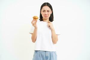 portret van ongelukkig Koreaans vrouw looks Bij koekje met verdrietig gezicht, meisje Aan eetpatroon kan niet eten gebakje, concept van gezond voedsel, wit achtergrond foto