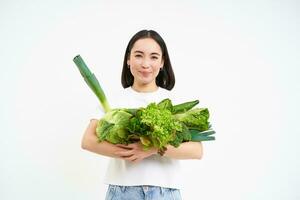 beeld van glimlachen Aziatisch vrouw met groenten, aan het eten gezond groen biologisch voedsel, geïsoleerd Aan wit achtergrond foto