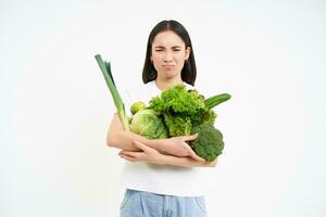 ongelukkig Koreaans vrouw, Holding groenten, gevoel ongelukkig Aan eetpatroon, aan het eten rauw groen voedsel, wit achtergrond foto