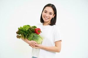 portret van mooi glimlachen Koreaans vrouw, houdt boeket van groenten, buying groen biologisch voedsel Bij markt, wit achtergrond foto
