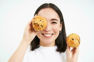 dichtbij omhoog portret van glimlachen Koreaans vrouw, shows smakelijk cupcakes, aan het eten taart en op zoek Vrolijk, gebakje en bakkerij concept foto