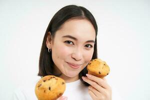 beeld van Koreaans vrouw geniet aan het eten gebakken gebakje, tonen twee smakelijk cupcakes in de buurt gezicht en glimlacht, wit achtergrond foto