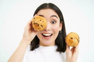 dichtbij omhoog portret van grappig Aziatisch meisje, houdt cupcakes in de buurt gezicht, maakt gelukkig gezichten, wit studio achtergrond foto