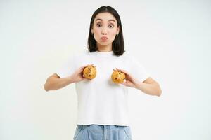 gelukkig Japans vrouw shows cupcakes Aan borst, aan het eten gebakje, staand over- wit achtergrond foto