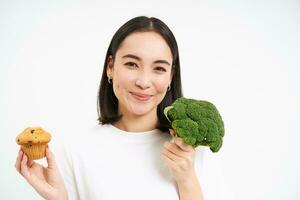 portret van Koreaans vrouw model, Holding koekje en broccoli, aan het eten gezond voedsel, kiezen mooi zo eetpatroon voor gezond lichaam, wit achtergrond foto