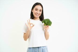 portret van gezond glimlachen vrouw, shows broccoli en duimen omhoog, beveelt aan plakken naar voedzaam eetpatroon met groenten, wit achtergrond foto