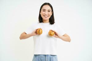 gelukkig Japans vrouw shows cupcakes Aan borst, aan het eten gebakje, staand over- wit achtergrond foto
