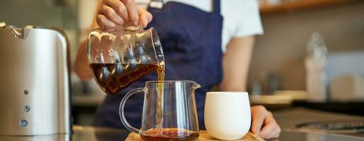 dichtbij omhoog van glas pot en een kop staand Aan cafe balie, barista gieten filter koffie en voorbereidingen treffen bestellen foto