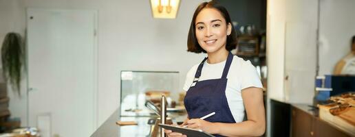 klein cafe bedrijf. glimlachen Aziatisch meisje barista in schort, gebruik makend van tablet net zo pos terminal, verwerken koffie bestellen achter teller foto