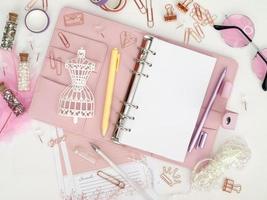 bovenaanzicht van een roze planner met schattig briefpapier. roze glamourplanner met een wit paspopbeeldje. planner met open pagina's op een witte achtergrond en met mooie accessoires pennen, knopen, pinnen. foto