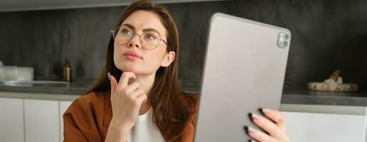 dichtbij omhoog portret van echt vrouw werken van huis, Holding digitaal tablet, op zoek terzijde met denken gezicht, nadenken, aan het studeren online, werken Aan afgelegen foto