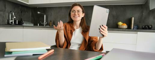 portret van jong eigen baas vrouw, zittend Bij huis, gemaakt keuken haar werkplek, gebruik makend van digitaal tablet naar praten, doet mee online Cursus of vergadering foto