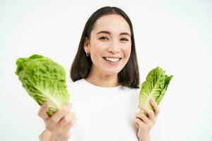 glimlachen Aziatisch vrouw, veganistisch aan het eten kool, geniet smakelijk vers sla bladeren, staat over- wit achtergrond foto
