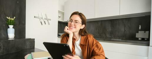 e-learning en afgelegen werkplaats concept. jong vrouw in bril, leerling aan het studeren Bij huis in keuken, lezing Aan digitaal tablet, op zoek Bij haar projecteren, verbindt naar online vergadering foto