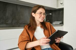 portret van jong leerling, vrouw aan het studeren Bij huis, werken van een afstand, opstelling werkplaats in haar keuken, zittend Aan stoel met digitaal tablet, lezing in bril foto