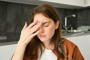 dichtbij omhoog portret van brunette vrouw aanraken hoofd, gevoel onwel, heeft hoofdpijn of migraine, zittend in keuken met duizeligheid. foto