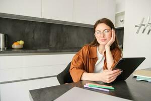 portret van jong leerling, vrouw aan het studeren Bij huis, werken van een afstand, opstelling werkplaats in haar keuken, zittend Aan stoel met digitaal tablet, lezing in bril foto