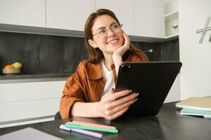 portret van jong leerling, vrouw aan het studeren Bij huis, werken van een afstand, opstelling werkplaats in haar keuken, zittend Aan stoel met digitaal tablet, lezing in bril foto