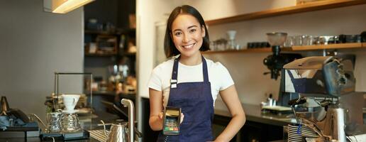 portret van Aziatisch barista meisje Bij balie, tonen kaart machine naar cliënt wie wil naar betalen contactloos, nemen bestellen, staand in cafe foto