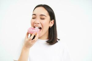 voedsel en aan het eten uit. jong Koreaans vrouw ziet eruit, duurt beet van heerlijk roze geglazuurd donut, wit achtergrond foto