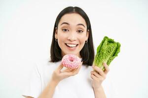 gezond schoon ontgiften aan het eten concept. vegetarisch, veganistisch, rauw concept. glimlachen Koreaans meisje eet heerlijk donut, houdt kool, geniet rommel voedsel, wit achtergrond foto