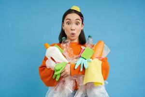 portret van Koreaans vrouw met verrast gezicht, Holding leeg plastic flessen en uitschot voor recyclen, blauw achtergrond foto