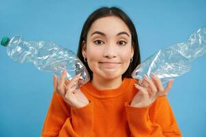 schattig Koreaans vrouw shows twee plastic flessen en glimlacht, recycleert, sorteert afval, verzamelen recyclebaar artikelen, blauw achtergrond foto