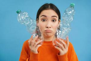 portret van verdrietig Aziatisch vrouw met plastic flessen, van streek door gebrek van recycling centra, blauw achtergrond foto