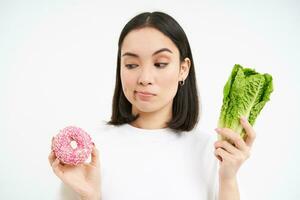 gezond levensstijl concept. jong vrouw beslissen tussen gezond groenten, kool en heerlijk geglazuurd donuts, wit achtergrond foto