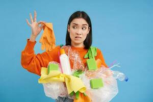 jong vrouw houdt plastic afval, looks Bij handschoen met geschokt gezicht, stinkend afval, sorteren afval voor recyclen, blauw achtergrond foto