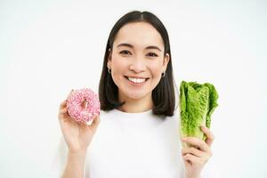 gezond levensstijl concept. glimlachen Aziatisch vrouw shows donut en kool, keuze van aan het eten ongezond rommel voedsel of groenten, wit achtergrond foto