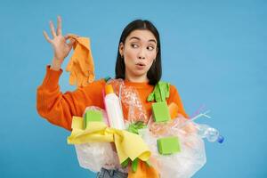 jong Aziatisch vrouw houdt afval, plastic flessen en afval, looks verrast Bij vuil latex handschoen, blauw achtergrond foto